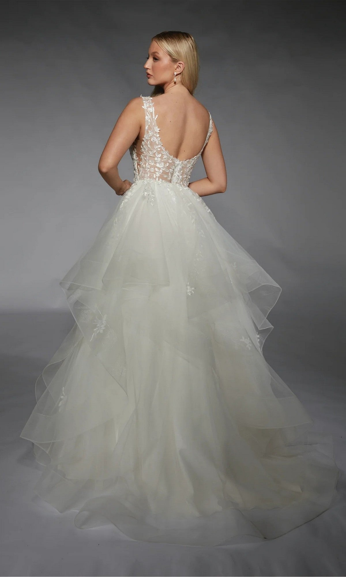 A woman faces away from the camera wearing the Long White Dress: Alyce 7126, an ivory ball gown with a sleeveless layered tulle skirt, sheer lace back, and delicate floral appliqués.